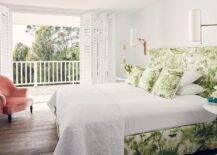 White and green bedroom features a green toile headboard on bed with matching pillows and bedskirt flanked by white round bedside tables illuminated by antique brass cylinder sconces facing a pink velvet armchair placed in front of plantation shutter folding doors leading out to a balcony.