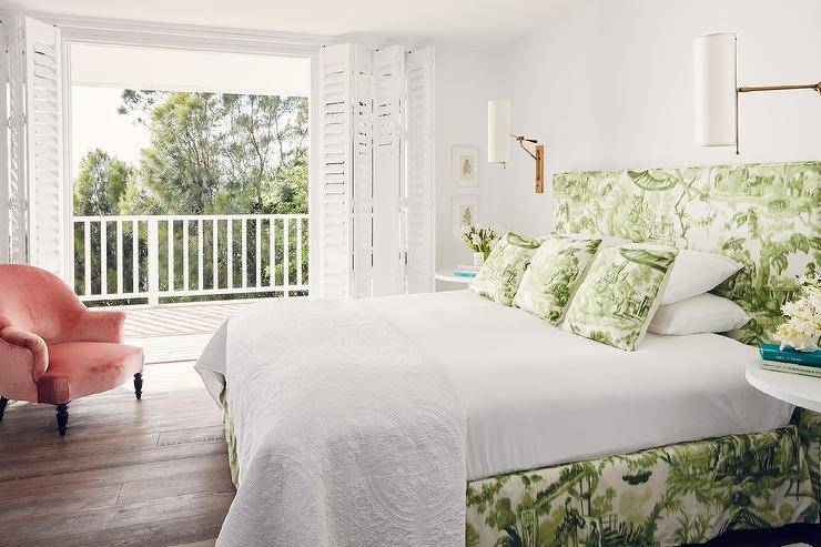 White and green bedroom features a green toile headboard on bed with matching pillows and bedskirt flanked by white round bedside tables illuminated by antique brass cylinder sconces facing a pink velvet armchair placed in front of plantation shutter folding doors leading out to a balcony.