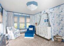 An Aerin Hampton Chandelier illuminates a blue nursery boasting a white campaign crib accented with white and blue toile curtains and positioned beneath a white and blue art piece hung from a wall clad in Nina Campbell Barbary Toile Wallpaper lined with blue crown molding. In front of windows dressed in white and blue curtains and placed on a leopard print rug, a blue velvet roll arm chair with a matching ottoman and a white and blue toile accent chair flank a gold leaf accent table.