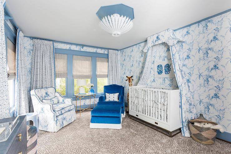 An Aerin Hampton Chandelier illuminates a blue nursery boasting a white campaign crib accented with white and blue toile curtains and positioned beneath a white and blue art piece hung from a wall clad in Nina Campbell Barbary Toile Wallpaper lined with blue crown molding. In front of windows dressed in white and blue curtains and placed on a leopard print rug, a blue velvet roll arm chair with a matching ottoman and a white and blue toile accent chair flank a gold leaf accent table.