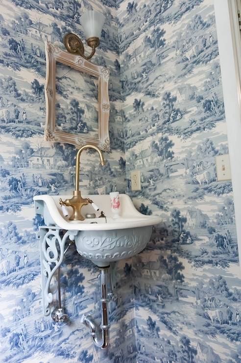 Incredible powder room with Sapphire Blue Toile wallpaper framing a distressed picture frame below a brass and frosted glass wall sconces. The ornate wall mounted sink with base and corbels painted in Benjamin Moore's Silver Lake pairs with a gooseneck brass faucet.