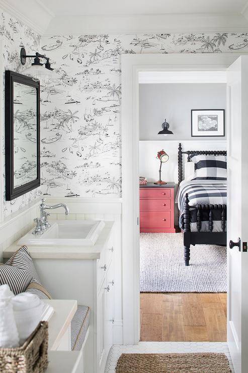 Cottage style kids bathroom with black and white toile wallpaper showcasing a bench between white washstands and a sink with vintage hook and spout faucets. Black inset framed medicine cabinets are mounted on the toile wallpaper finished with a two-light black sconce.