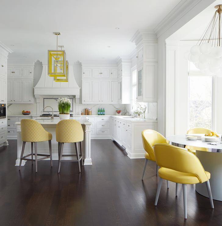 White and yellow kitchen colors create a bright, airy and cheerful setting. Here, yellow seating becomes a theme in the space complementing white cabinetry and walls. A set of yellow fabric counter stools are designed at a white island with a gooseneck sink illuminated by yellow lanterns. Yellow fabric counter stools at a round chrome trim kitchen table bring a pop of color to the white and silver tones at a bay window finished with a globe cluster chandelier.