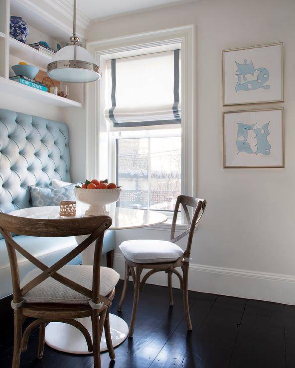 Chic white and blue keeping room features French x-back dining chairs placed at a round white tulip table facing a turquoise blue tufted banquette bench fitted beneath white shelves. An industrial pendant lights the table, as a gray grosgrain roman shade covers a window positioned beside stacked white and blue abstract art.