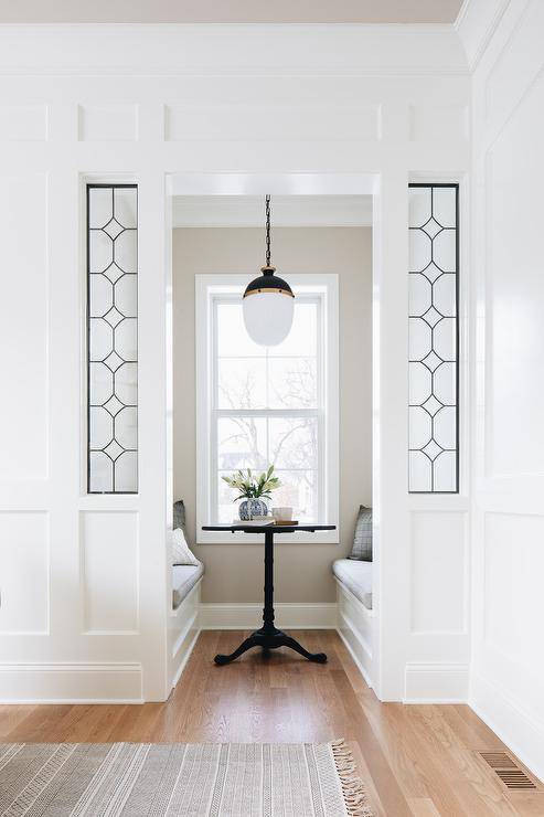 Peak through leaded glass panels into a breakfast nook boasting a black French table flanked by built-in white benches topped with gray cushions. A black and gold pendant hangs over the table and in front of an uncovered window framed by light gray wall paint.