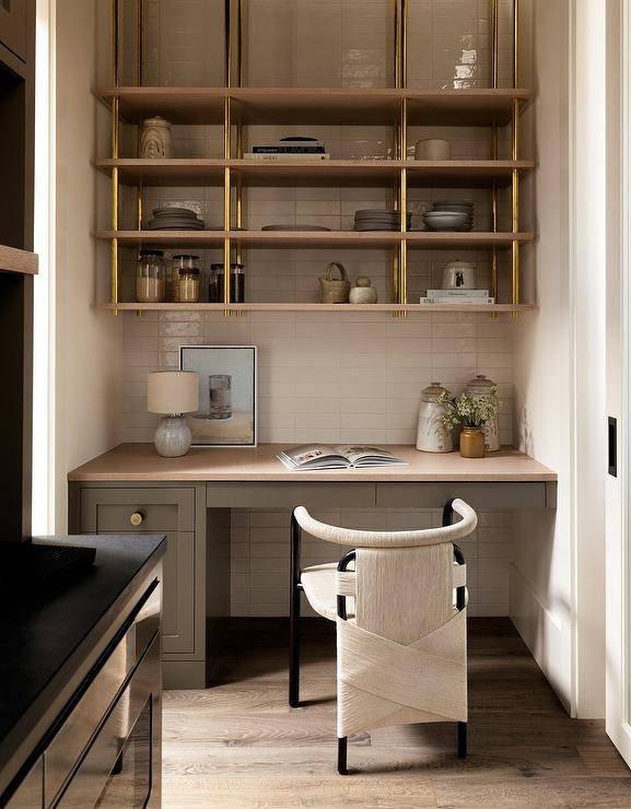 A small office space off kitchen features a black and white rope chair at a taupe built in desk under wood and brass shelves on white stacked backsplash tiles.