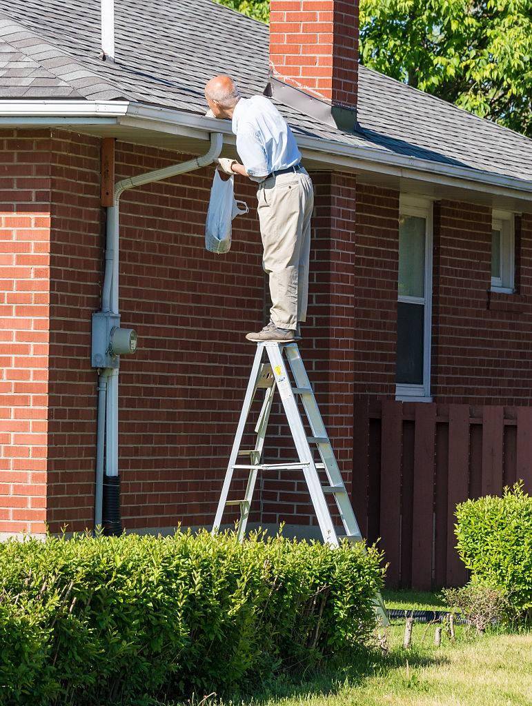 Senior man cleaning a rain gutter on a ladder. Clearing autumn gutter blocked with leaves by hand.