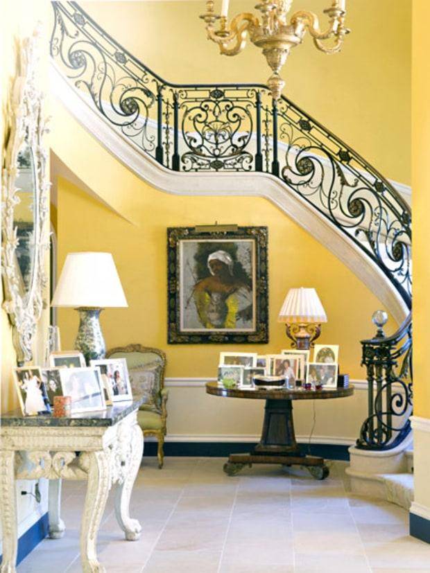 An entrance hall includes tables with personal photos, a yellow staircase, and a painting.