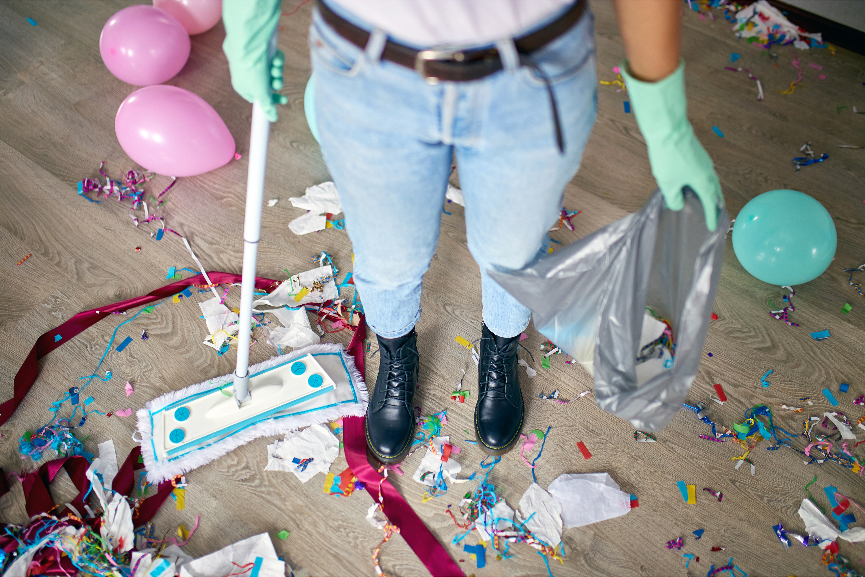 Looking down at the legs of a person holding a broom, a large party mess on the ground around them.