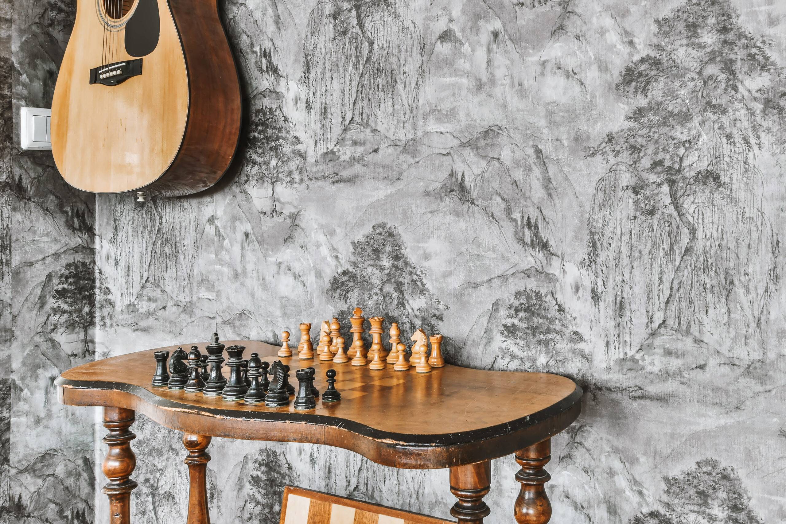 A table with a chess set beneath a hung up guitar, detailed wallpaper on the wall.