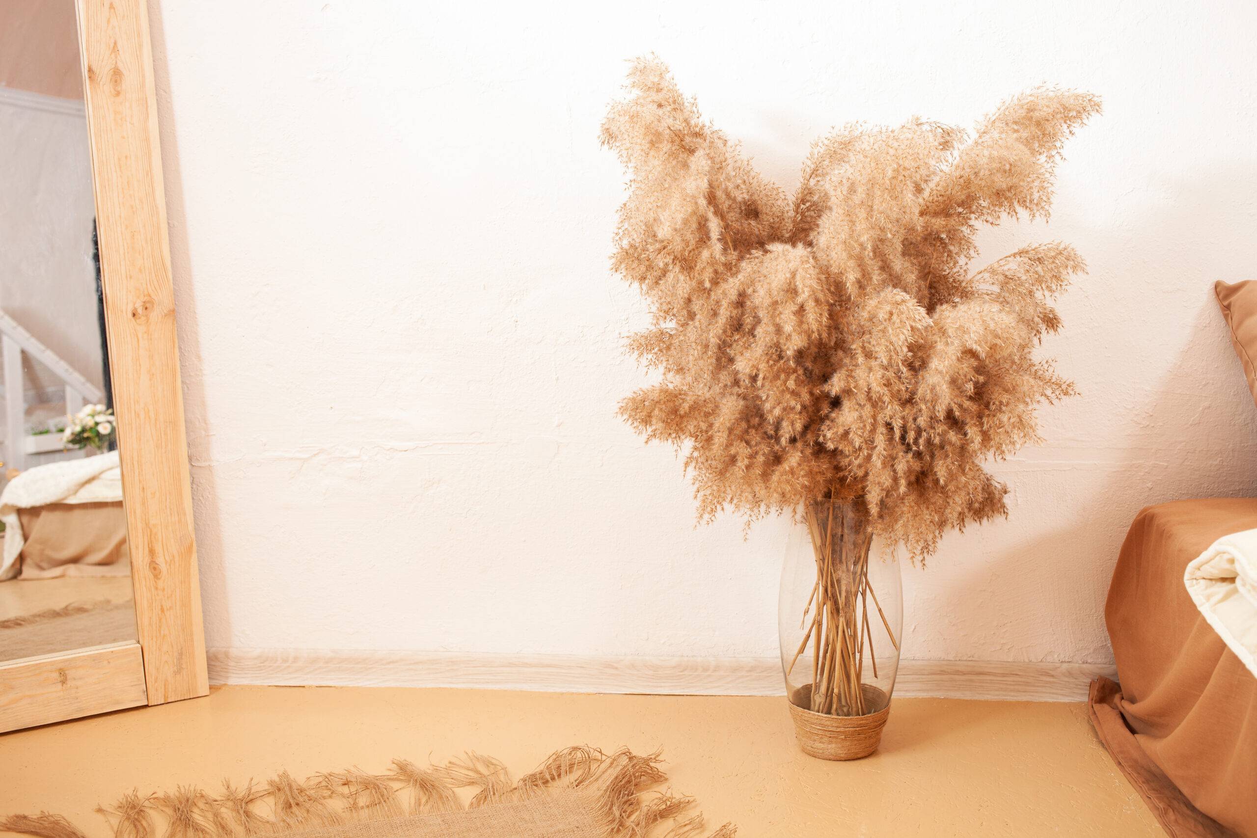 Pampas grass in a vase on the ground, a mirror and bed beside it.