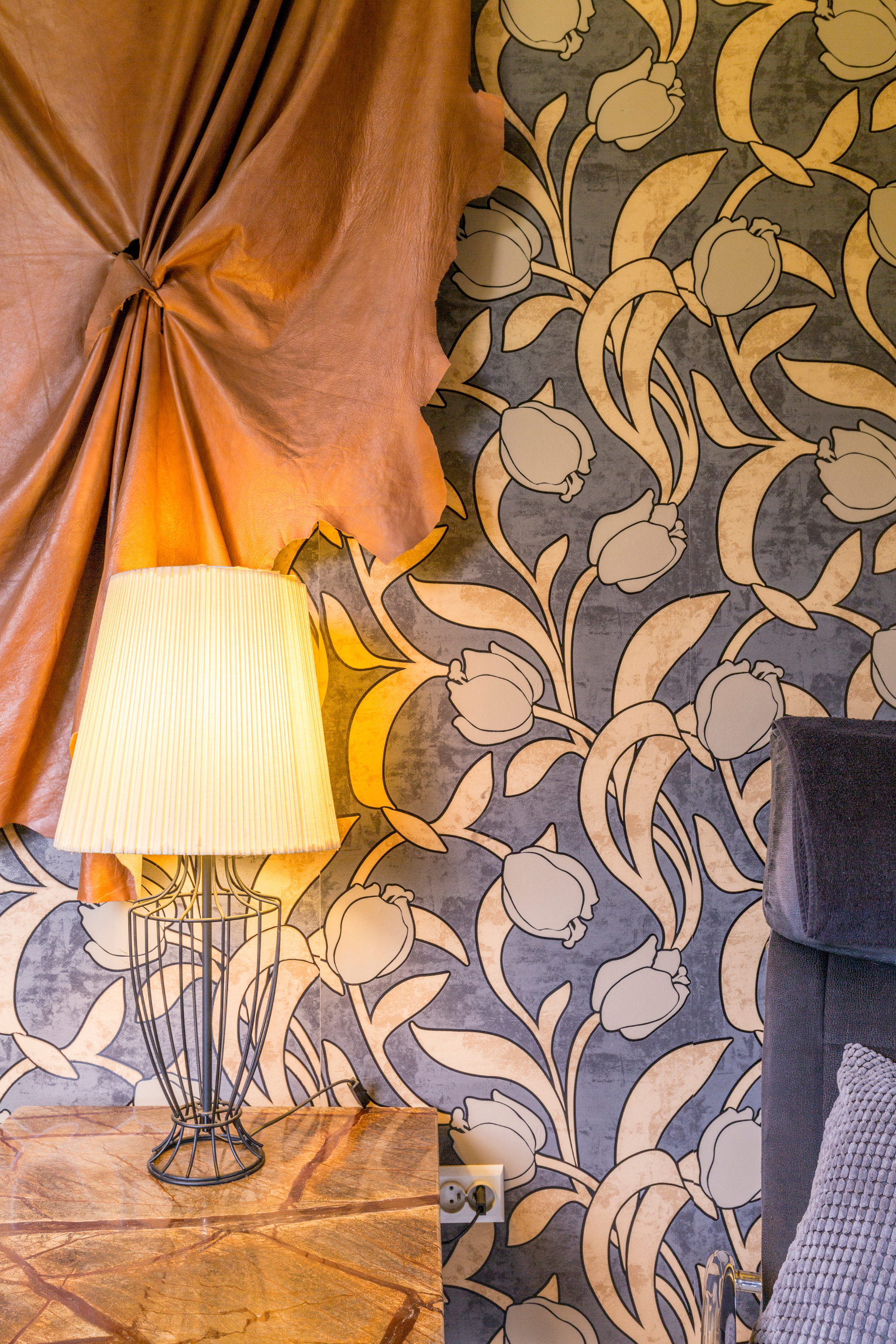 A beside table and a turned-on lamp, a curtain, and wallpaper on the wall.