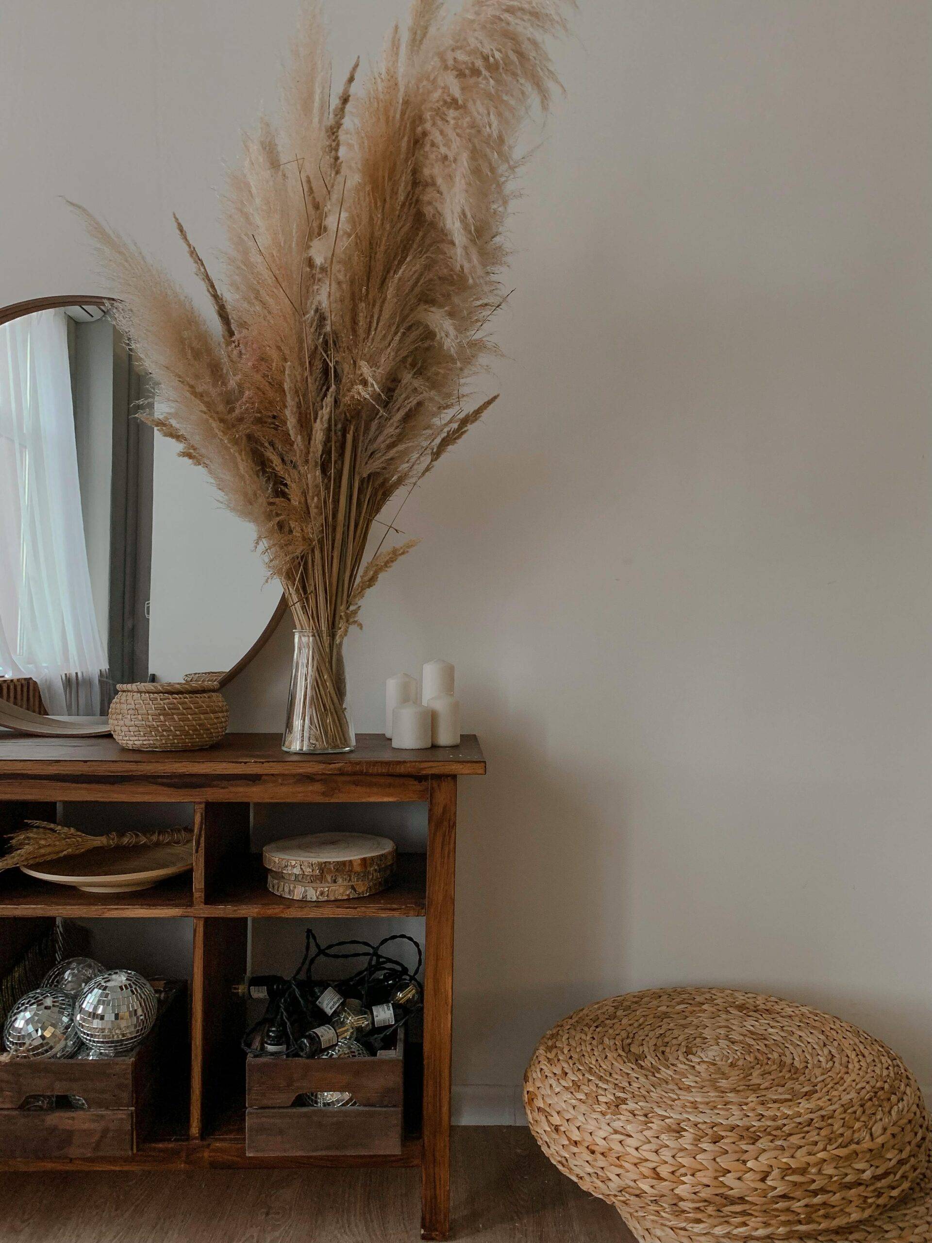 Pampas grass in a vase on a decorated dresser, an ottoman beside it.