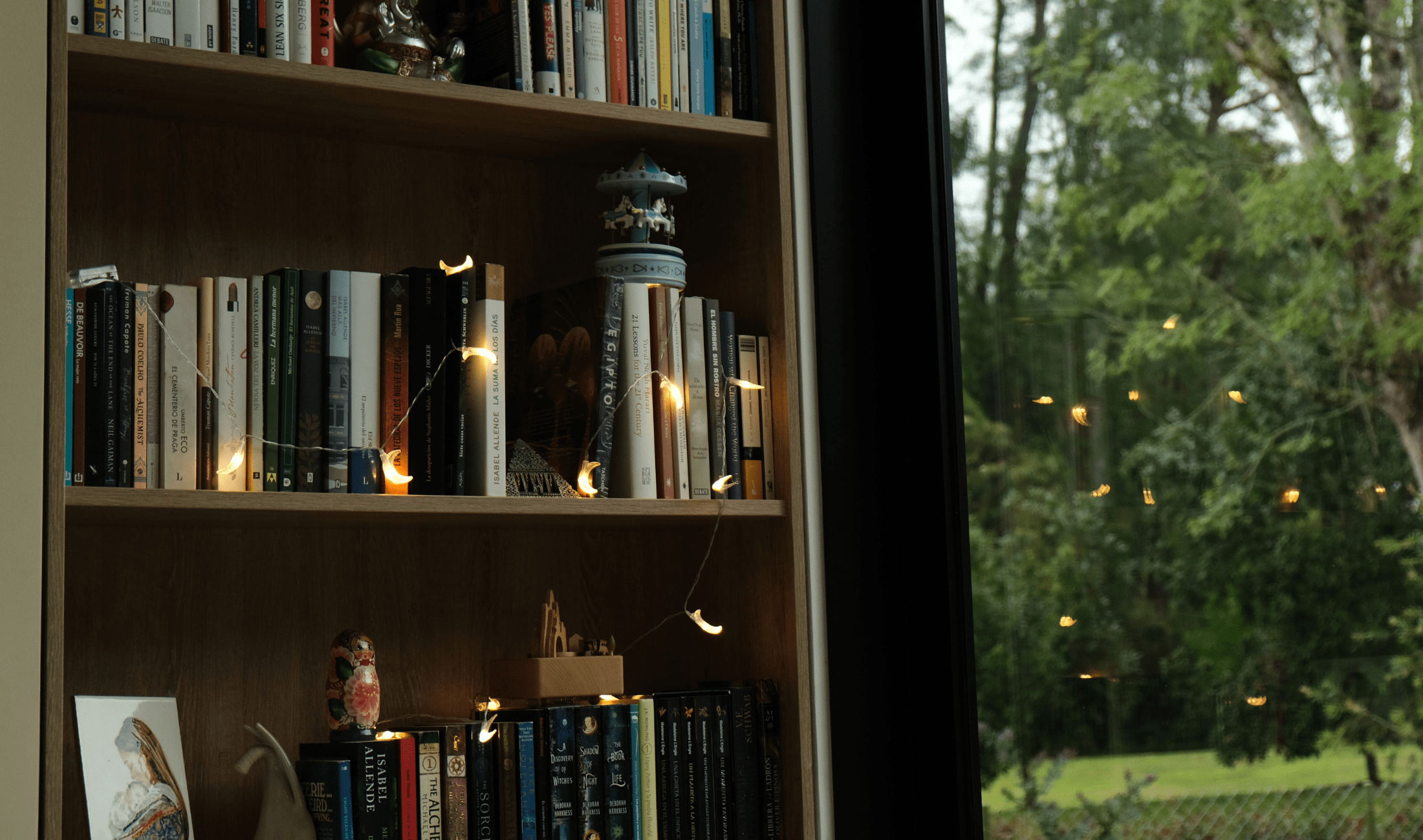 A cozy bookshelf adorned with fairy lights next to a window with rainy day outside.