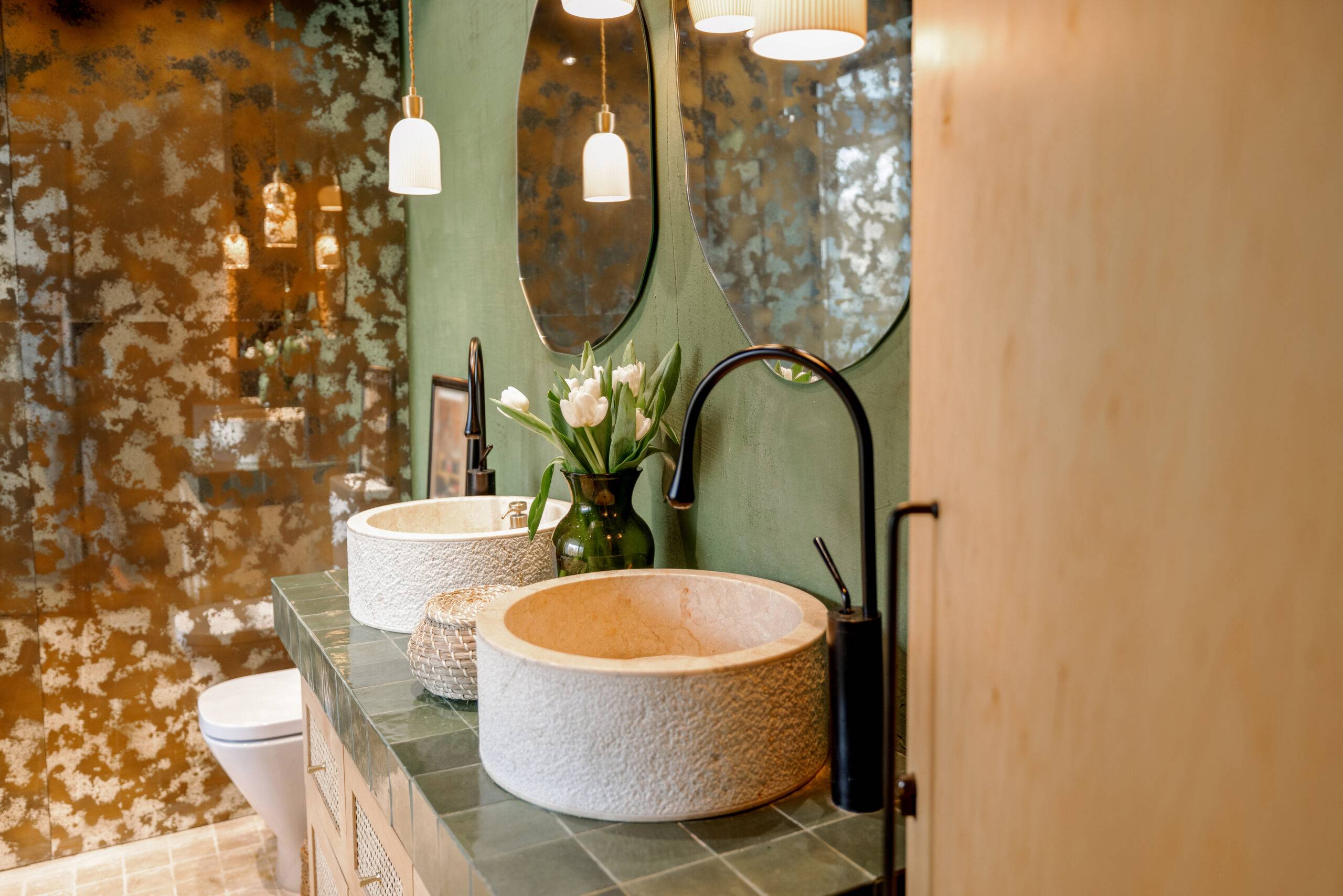 Bathroom interior with stone wash basins, modern faucets, irregularly shaped mirrors and wooden rattan commode in green and beige tones in natural boho style