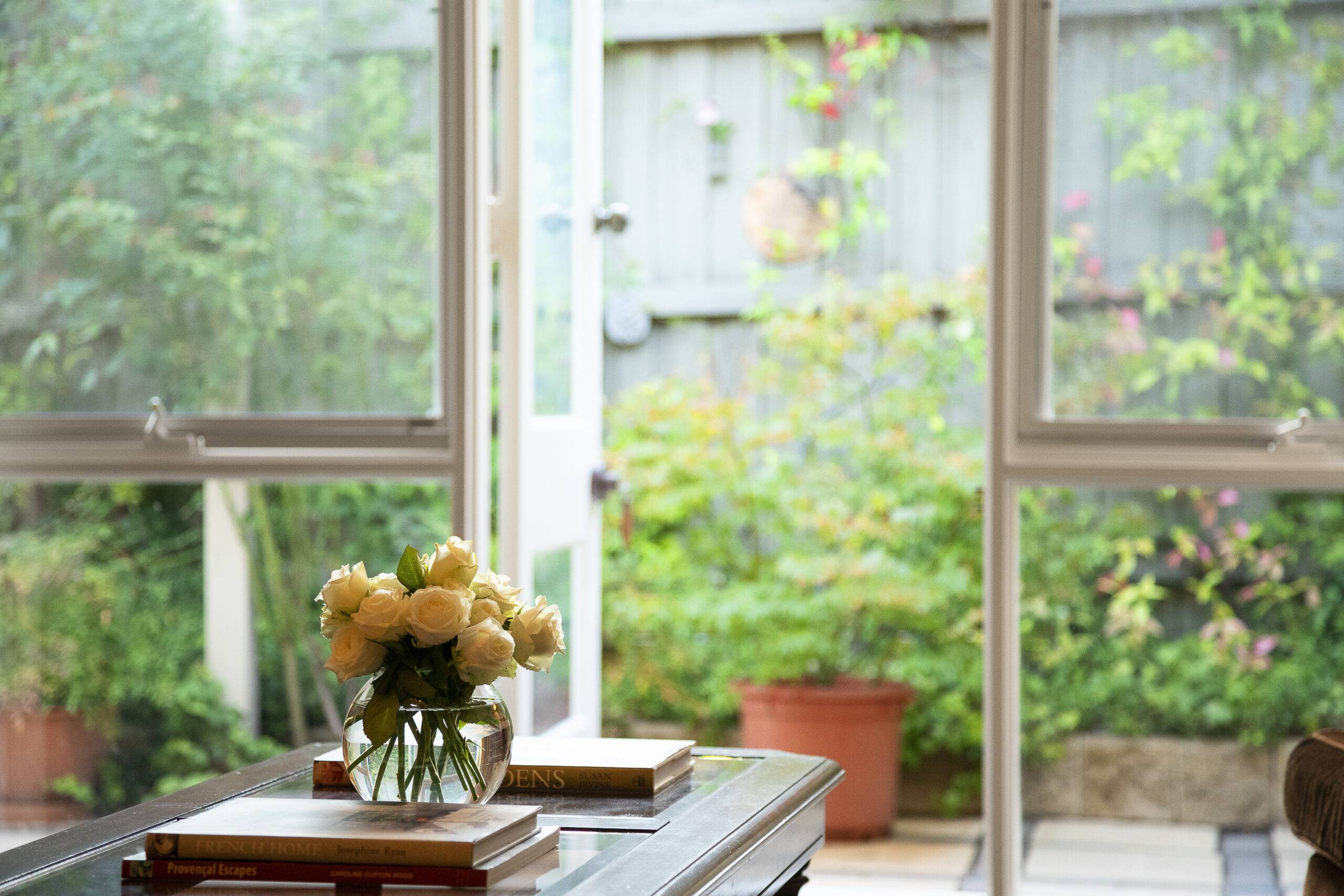 A table with a vase and a few books backing against large, full-wall window panels to a small outdoor courtyard.