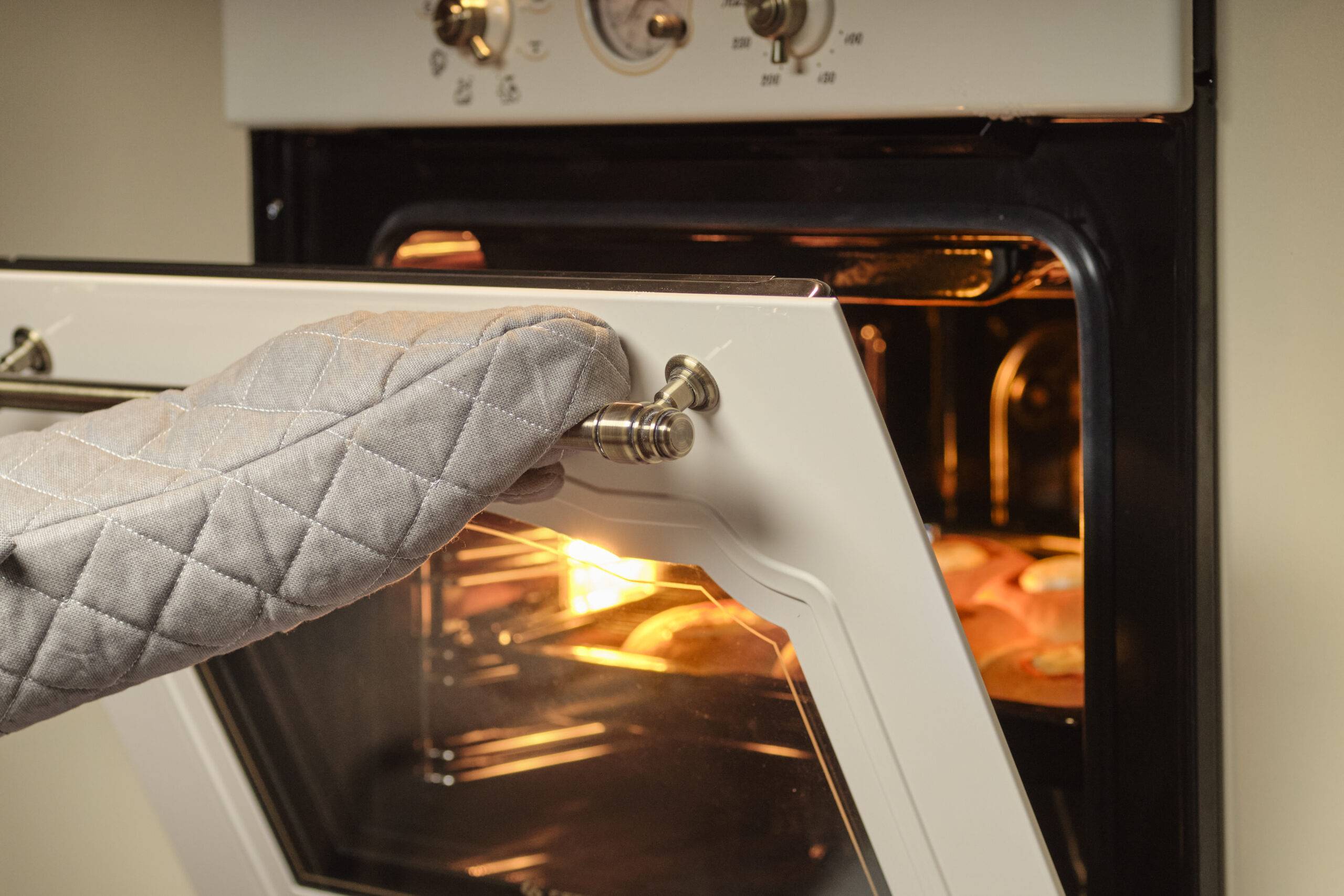 A hand wearing an oven mitt opens an oven door.