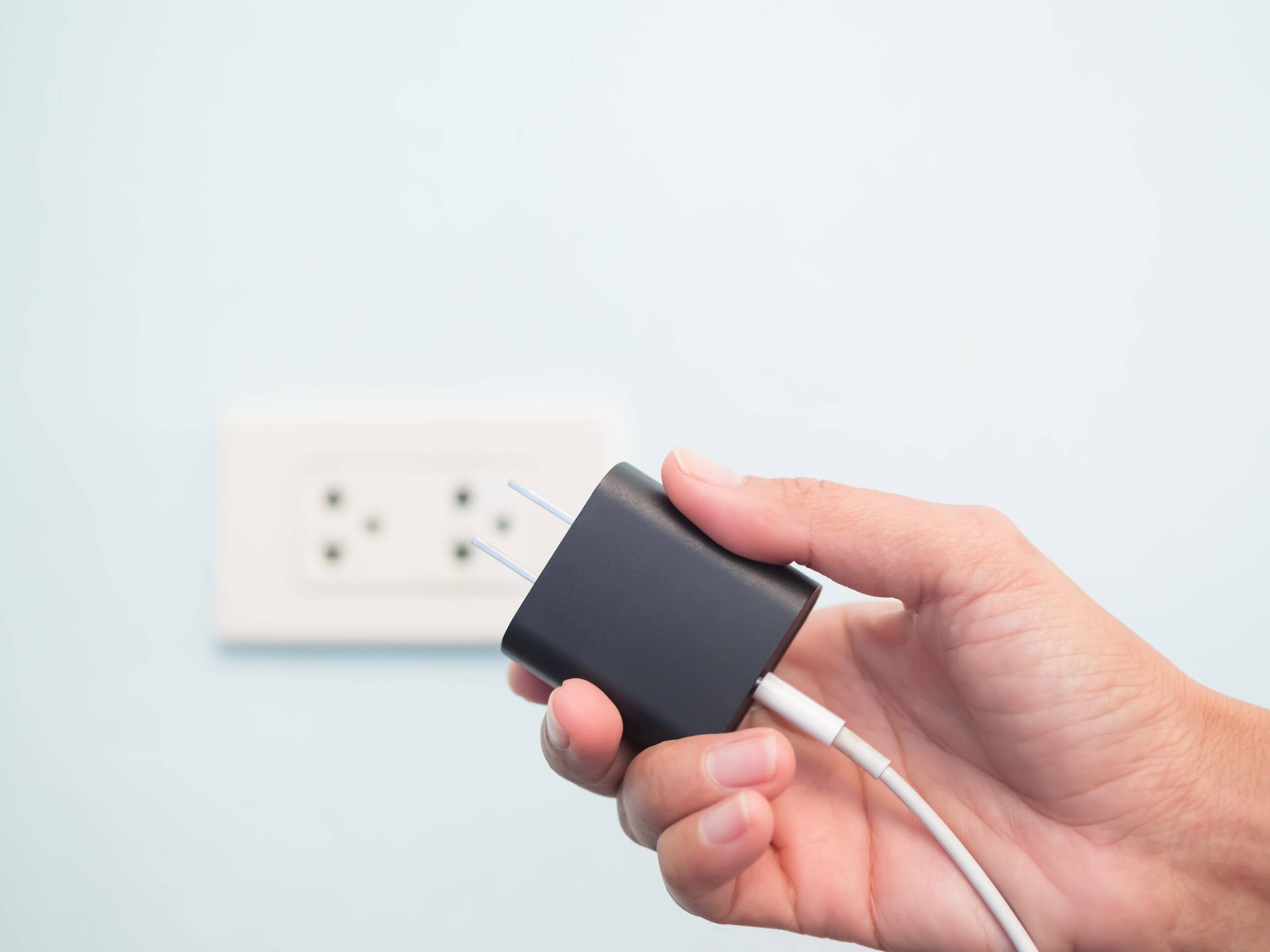 A hand holding a phone charging block in front of a wall outlet.