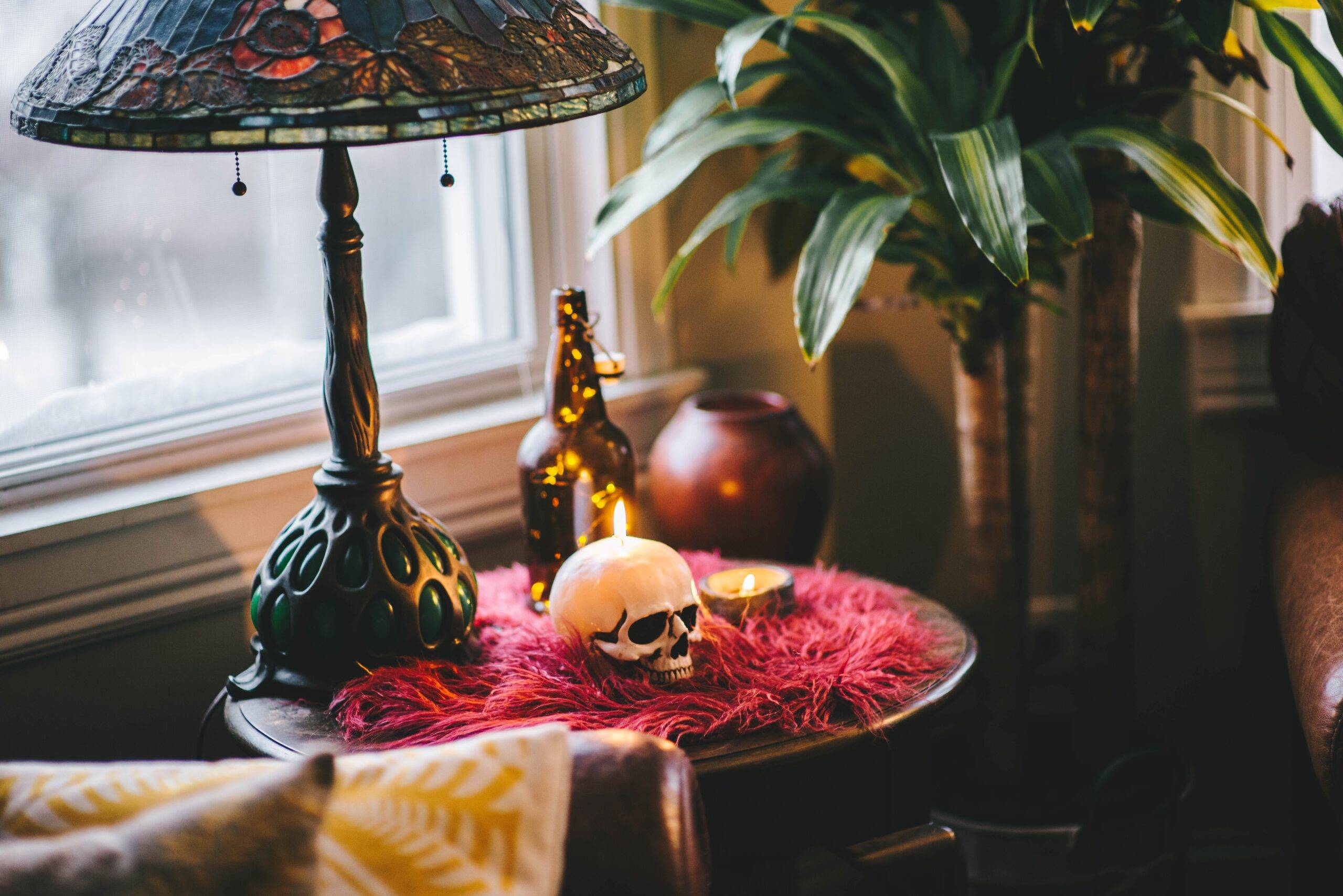 A side table in a dark, gothic home with a pink fuzzy cloth, a dark stained glass lamp, and a small decorative human skull.