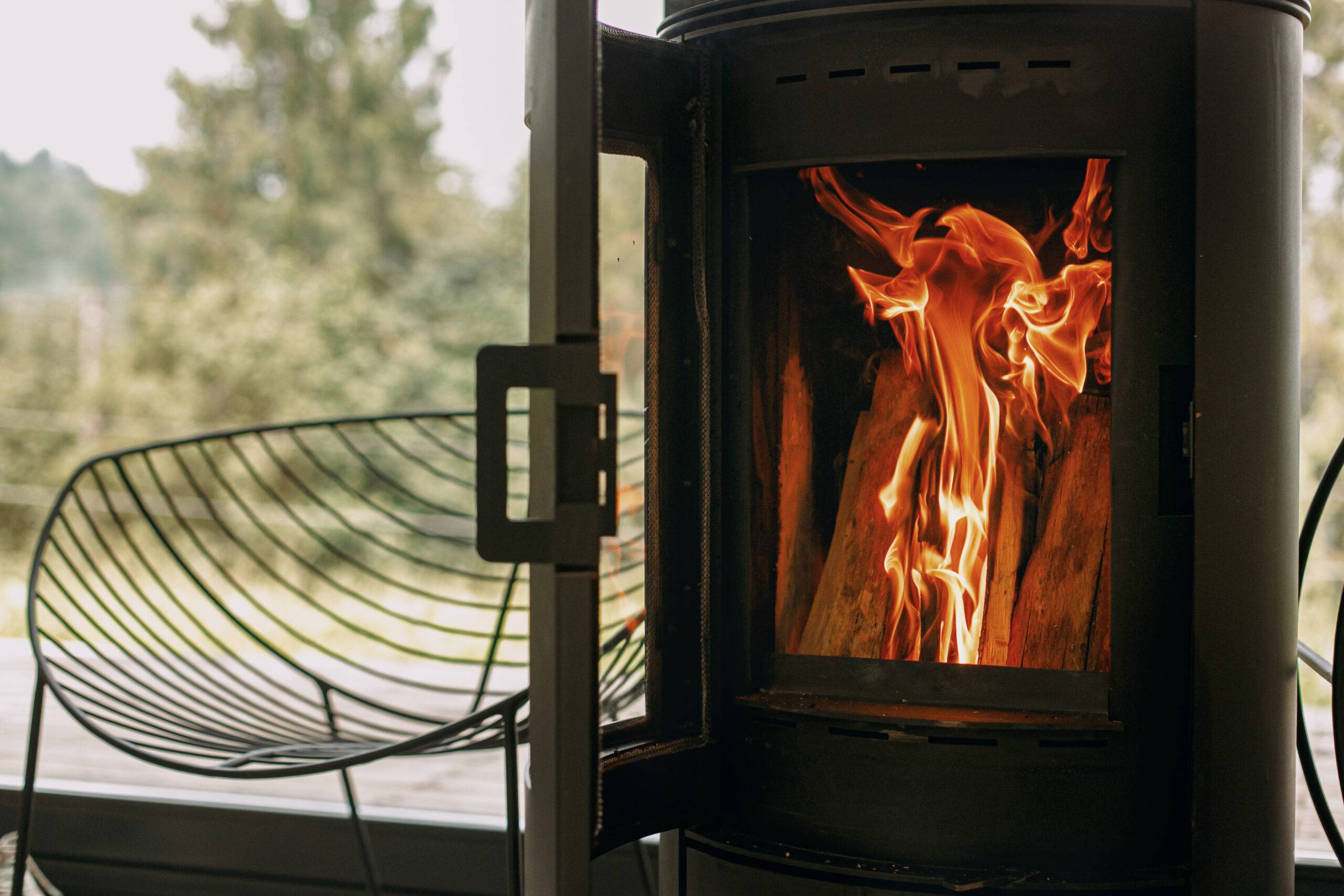 Modern black fireplace with burning fire and firewood on metal stand on background of big windows with view on mountains. Cozy warm and calm moments at cold season.