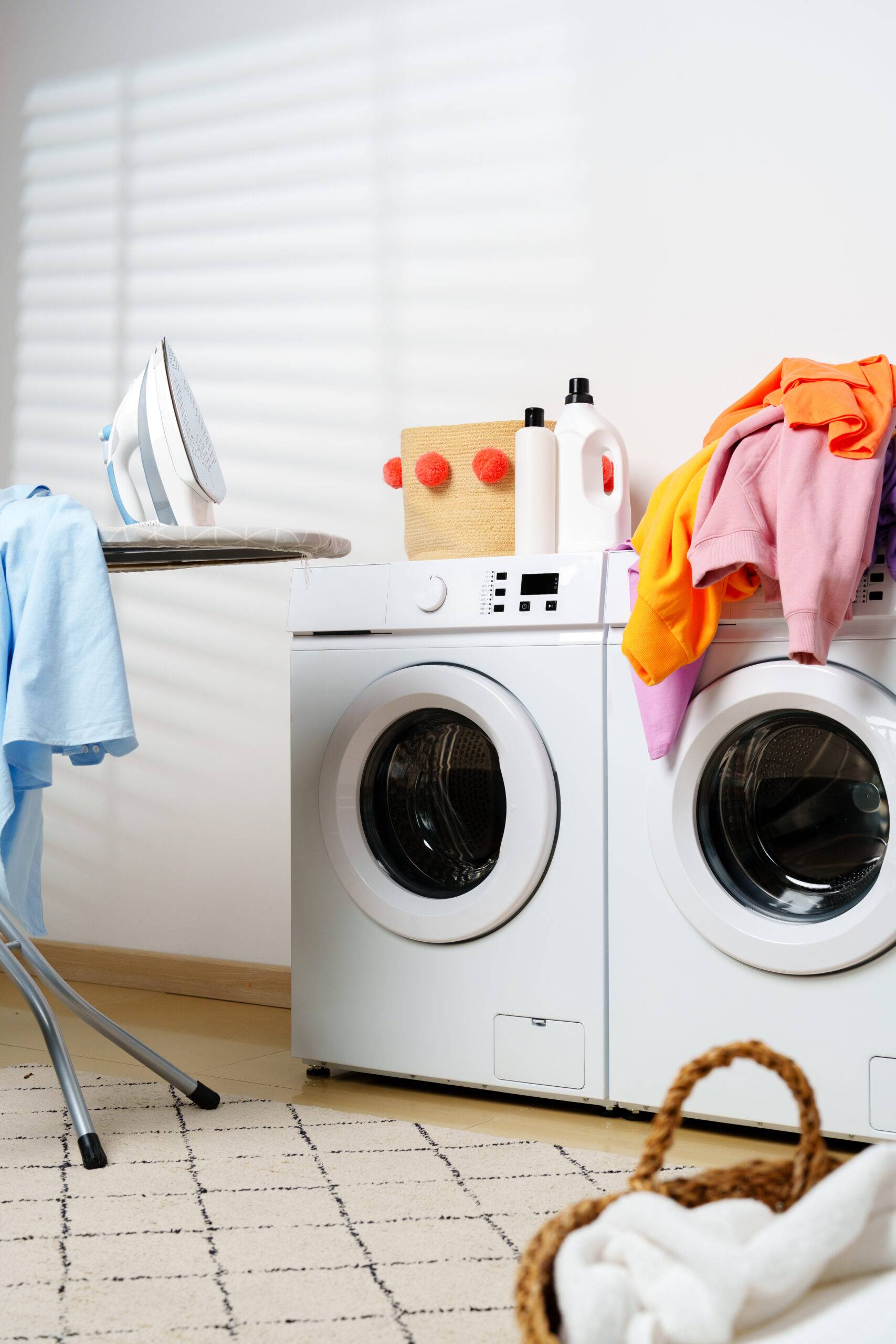 A washing machine and drying machine with clothes and detergent on top, an ironing board beside it.