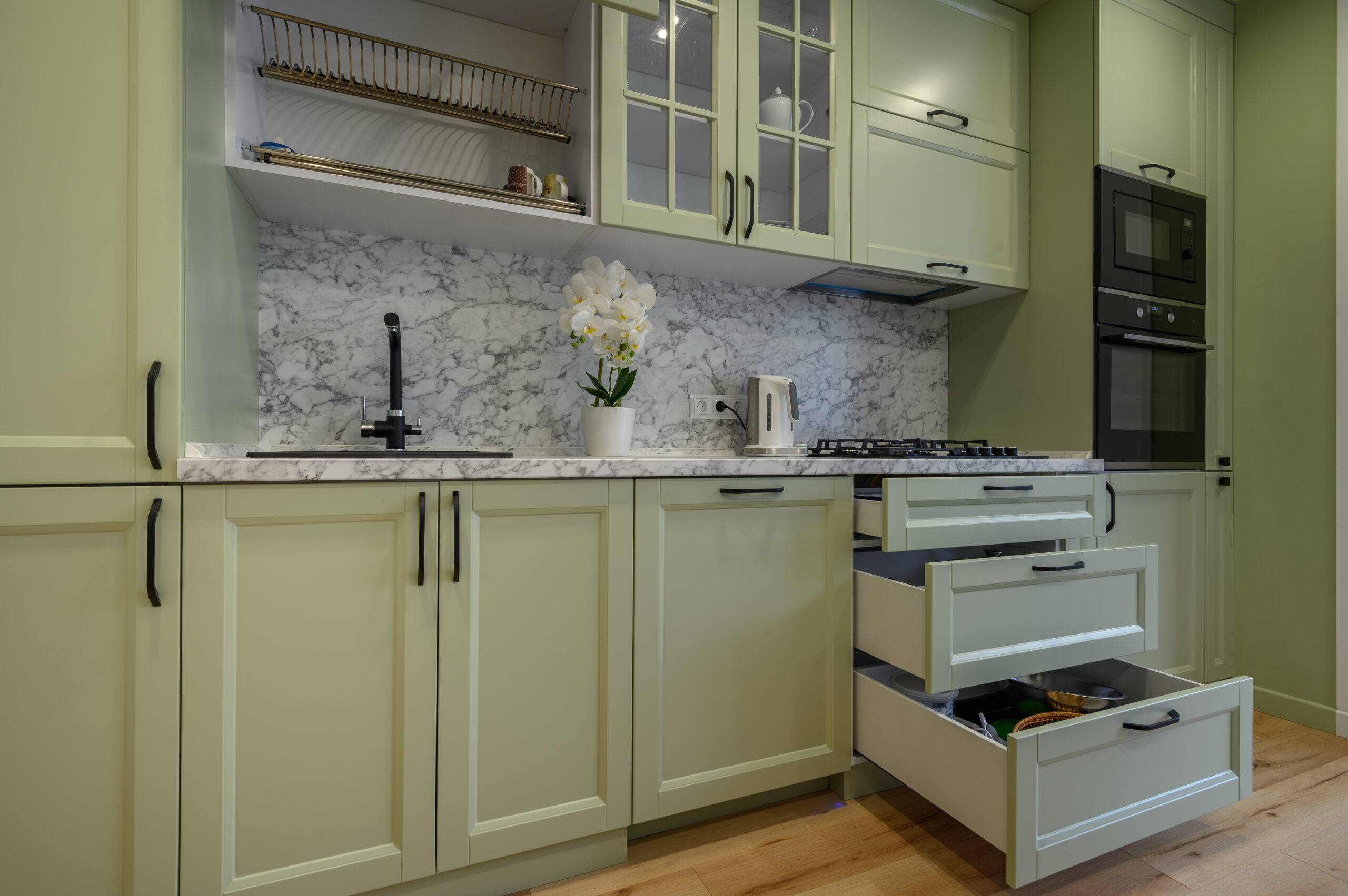 Modern trendy light green luxurious kitchen with dining table and dark green chairs, some drawers are retracted