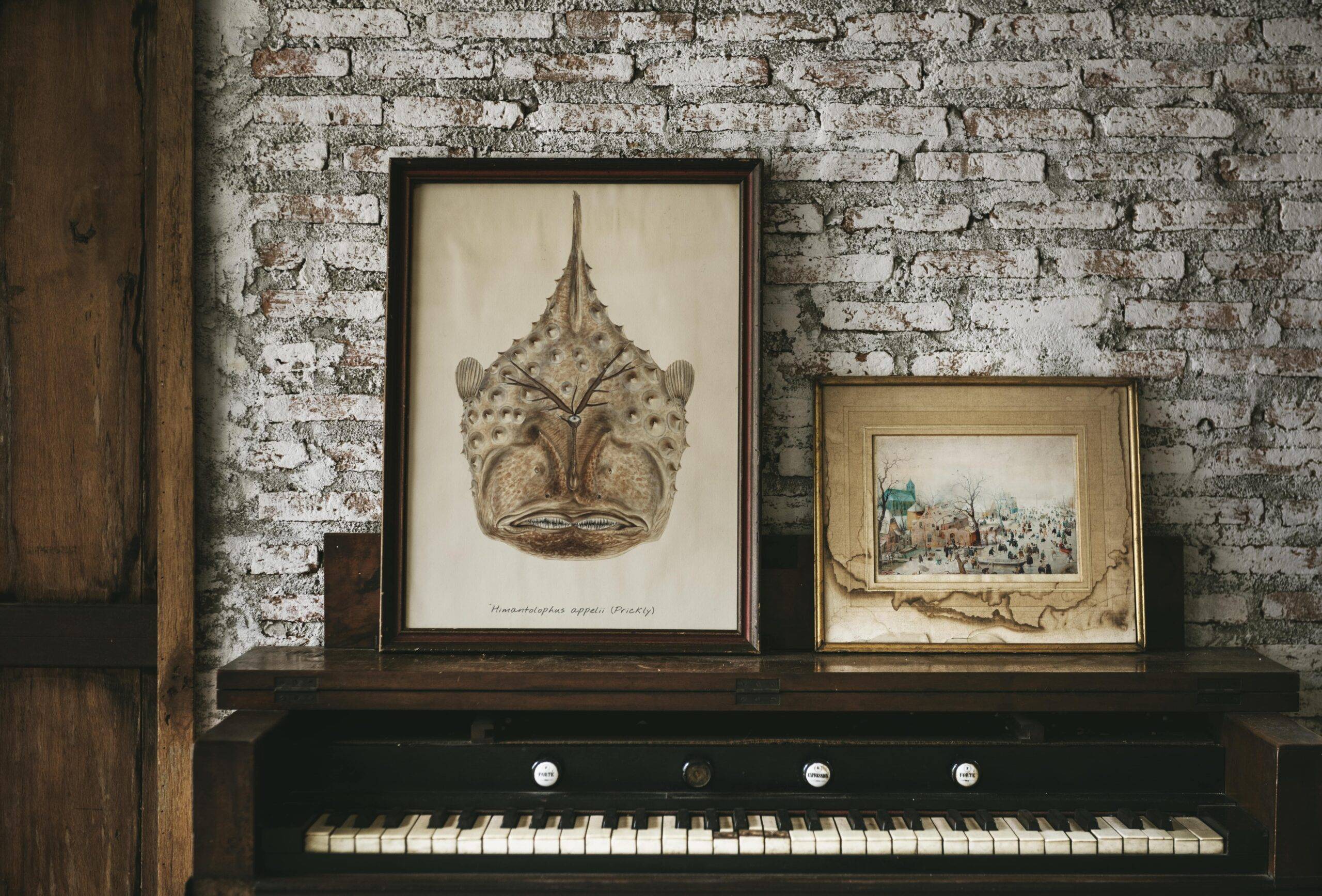 Two pieces of art in older frames resting on top of a piano against a brick wall.