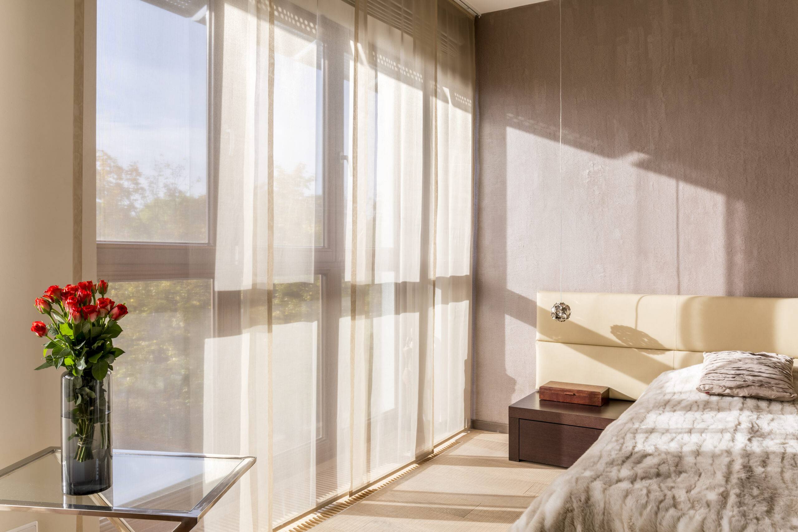 Spacious and elegant bedroom in beige colors with flowers and a bed, focus on the transparent curtains in the large window.