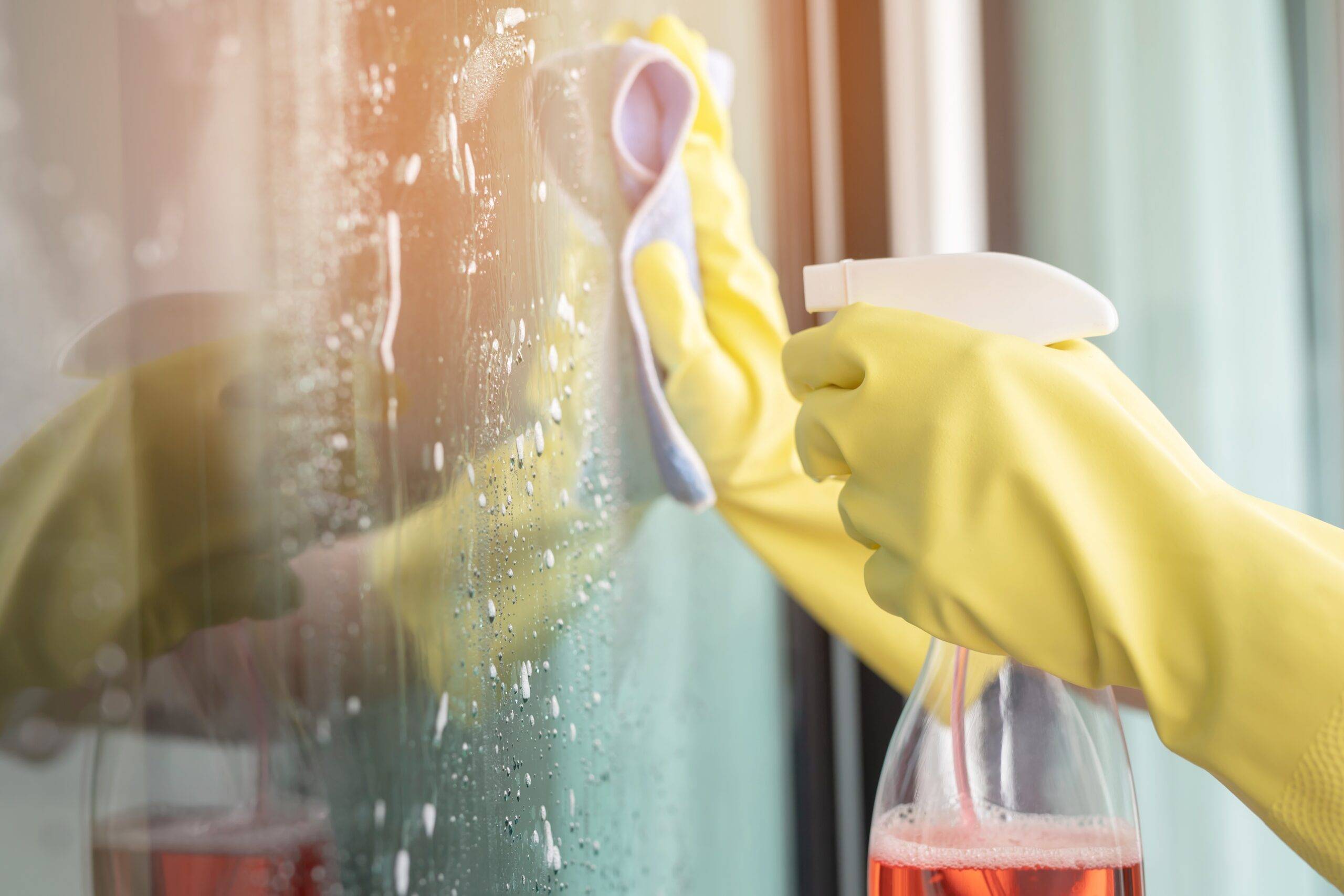 Someone in yellow gloves holding a spray bottle and rag, cleaning a window.