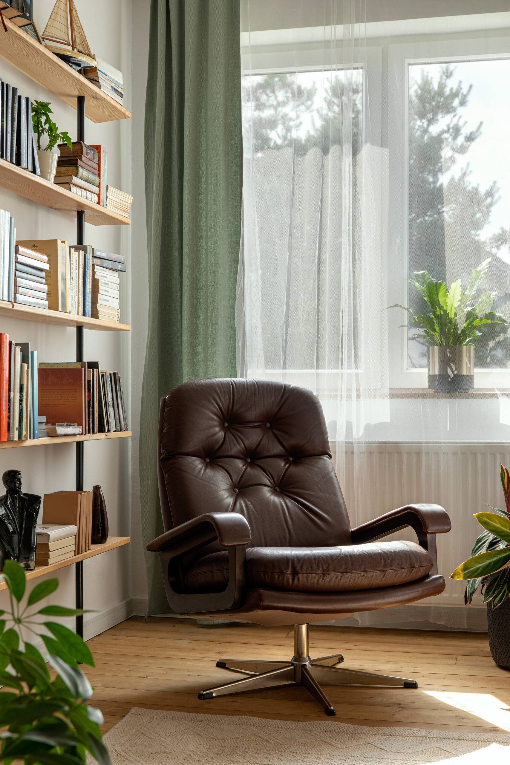 Stylish composition of home office interior with design retro armchair, library, plant, window, books, decoration and elegant personal accessories in home decor.