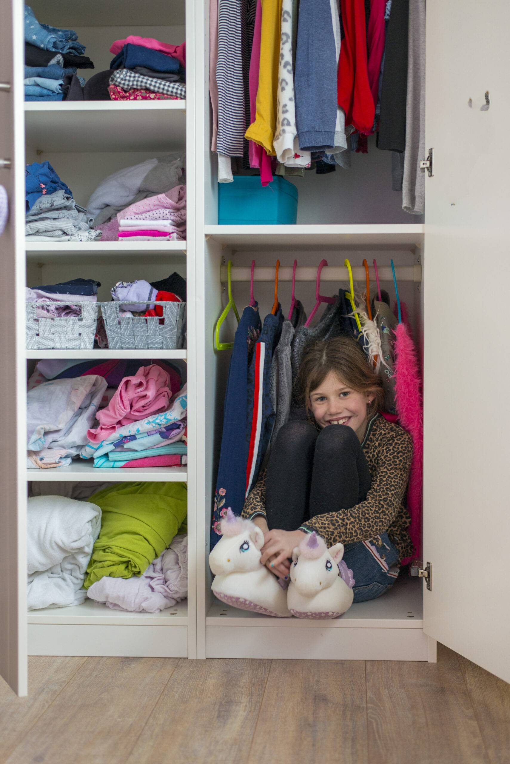 Young girl enjoying a playful moment hidden in a wardrobe full of clothes.