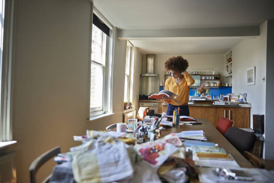 Young woman engrossed in reading a book in a cozy sunlit room filled with clutter.