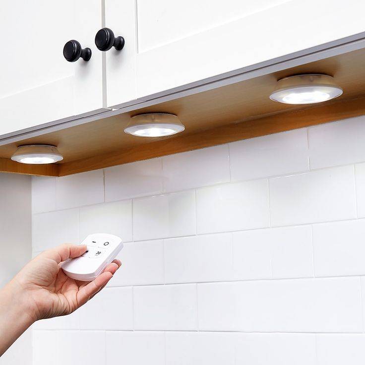 A set of puck lights under a cabinet, a hand reaching out to turn them on with a remote.