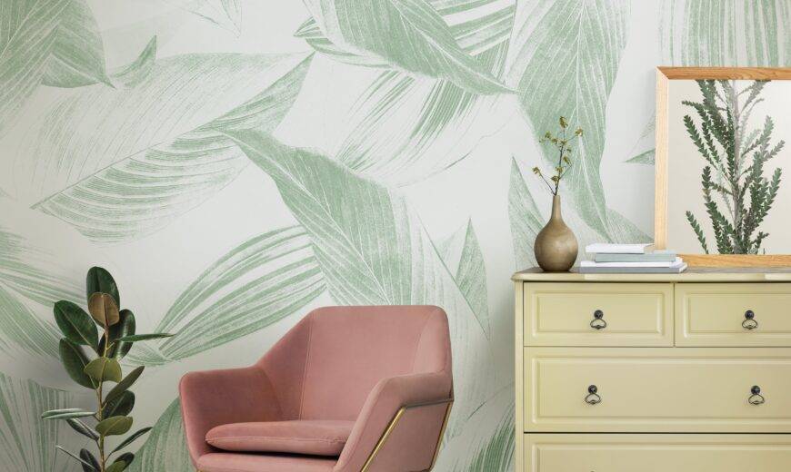 A section of a living room, zooming in on a chair and dresser with accessories, a large print leafy wallpaper behind.