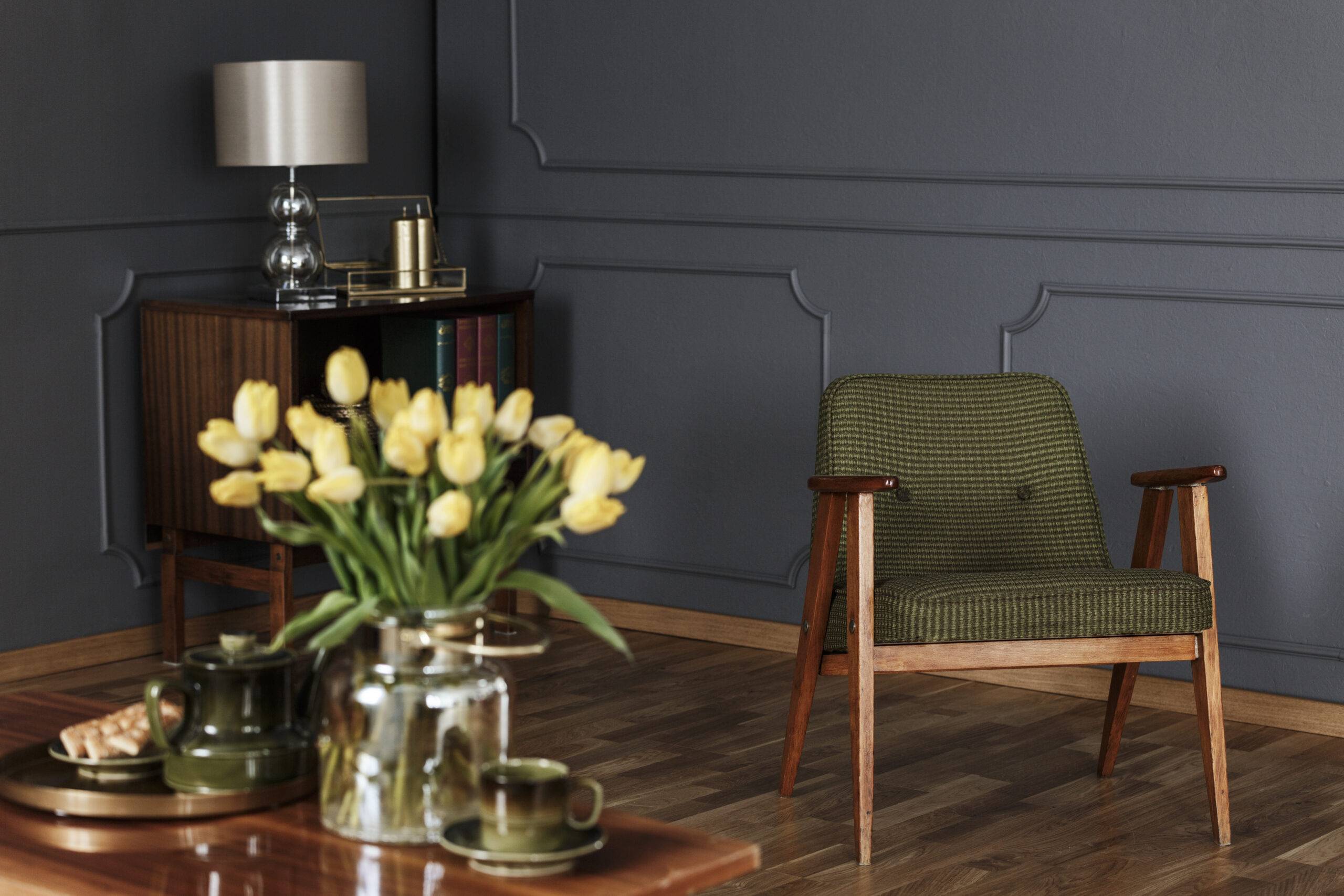 Real photo of a green, old-fashioned armchair in living room interior with a wooden cupboard in the background and blurred yellow tulips in the foreground