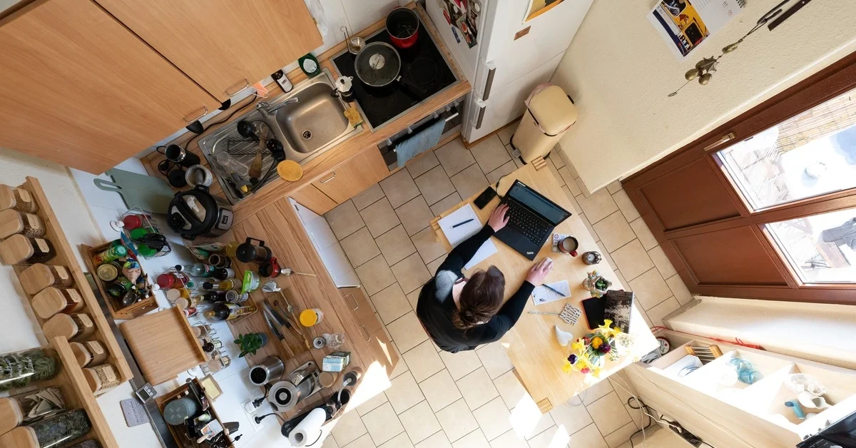 A woman sits at a table in a home office kitchen working on a laptop.